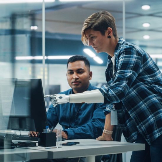 Teamwork In Diverse Inclusive Office: Woman with Disability Project Manager with Prosthetic Arm Talks with Indian Specialist Working on Desktop Computer. Professionals Software Engineers Create App